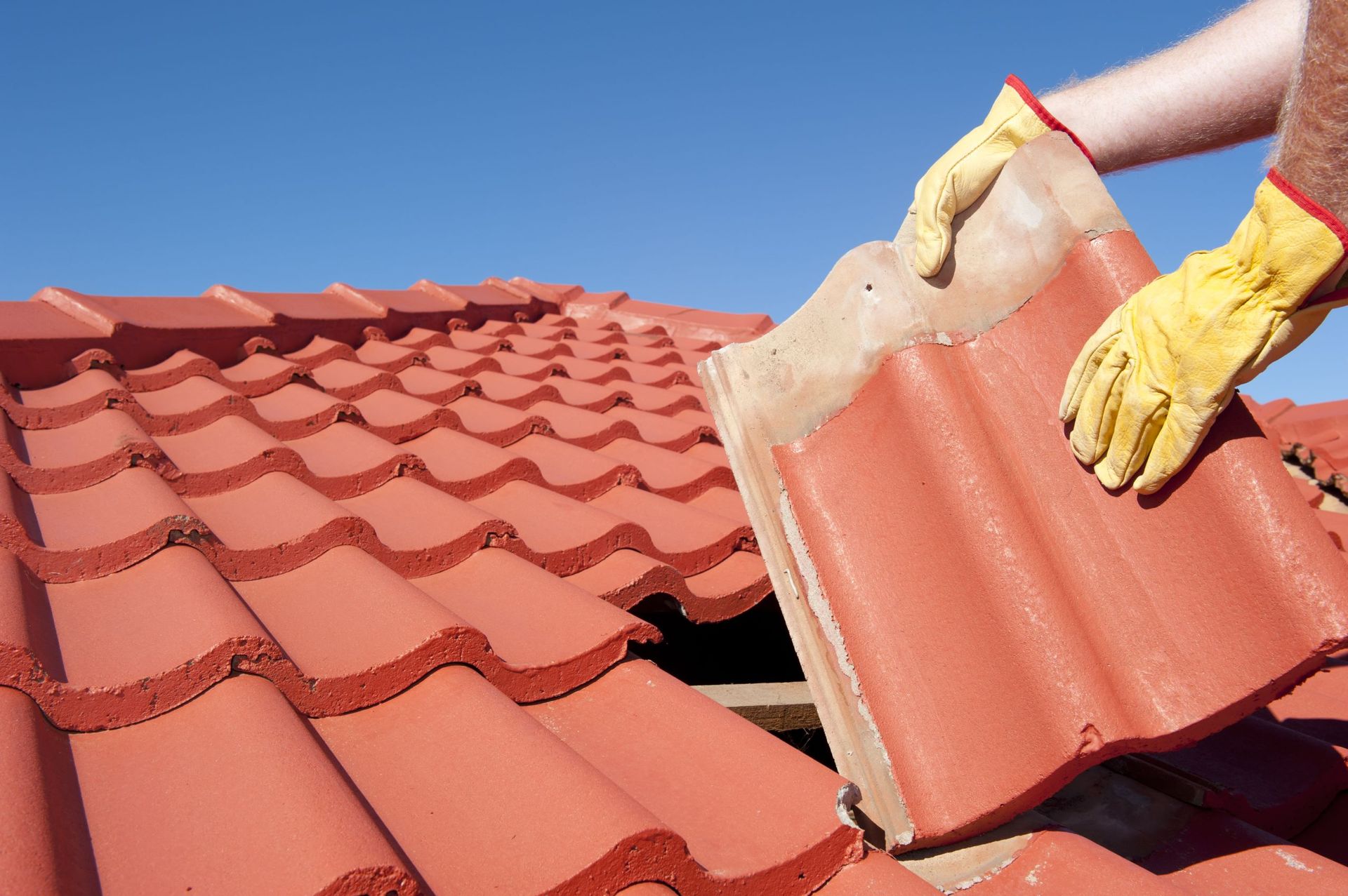 A person wearing yellow gloves is holding a red tile on top of a roof.