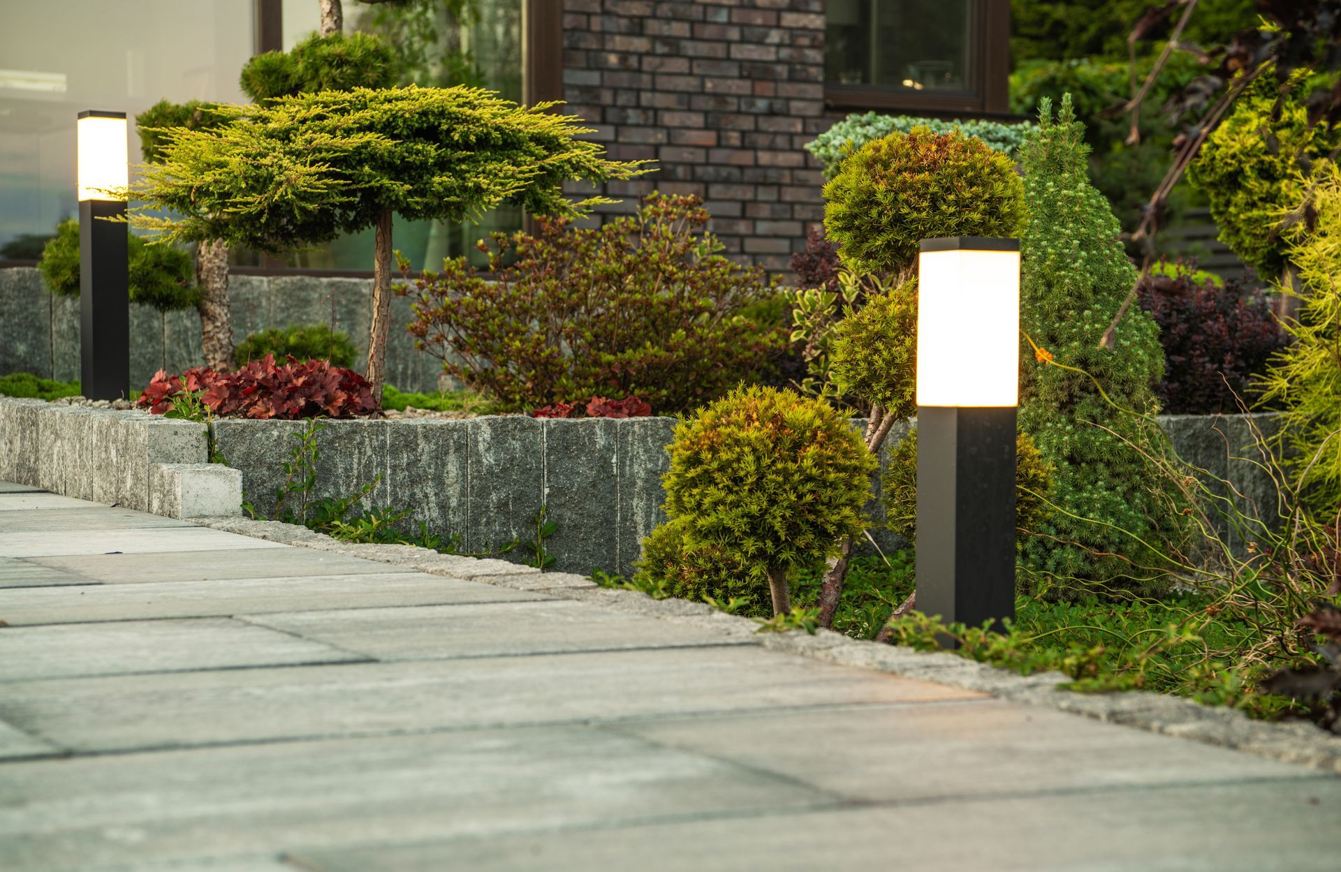 Outdoor Lights Installed Along Concrete Garden Path at the Residential House Backyard. Landscaped Flower Beds in the Background. Yard Illumination Theme.