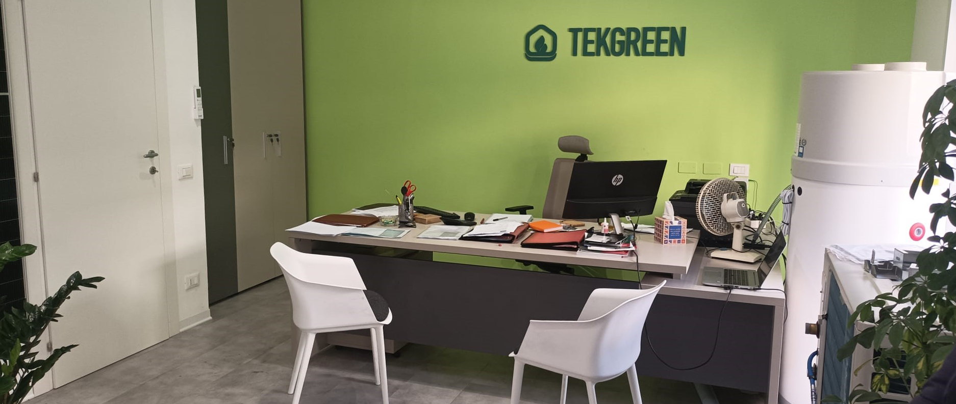 Ufficio con pareti verdi, scrivania, sedie, computer e piante in vaso sotto l'insegna TEKGREEN.
