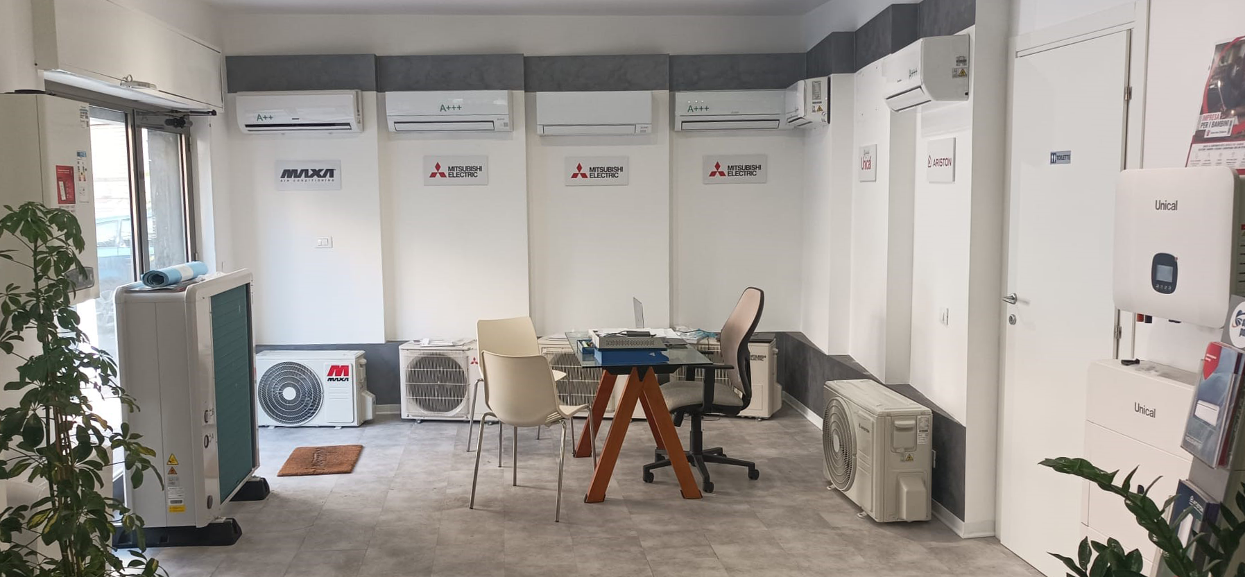 Stanza ingombro di sedie, tavolo, ventilatore, armadietti e piante lungo le pareti bianche.
