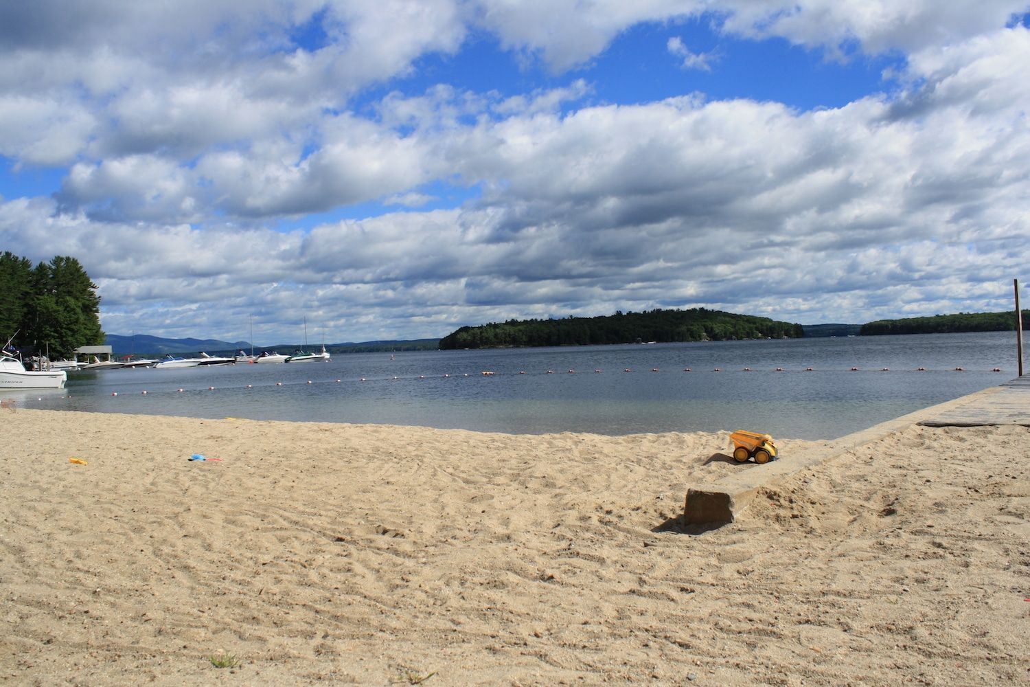 A sandy beach leading to a calm lake with a forested island in the distance under a blue sky with scattered clouds.