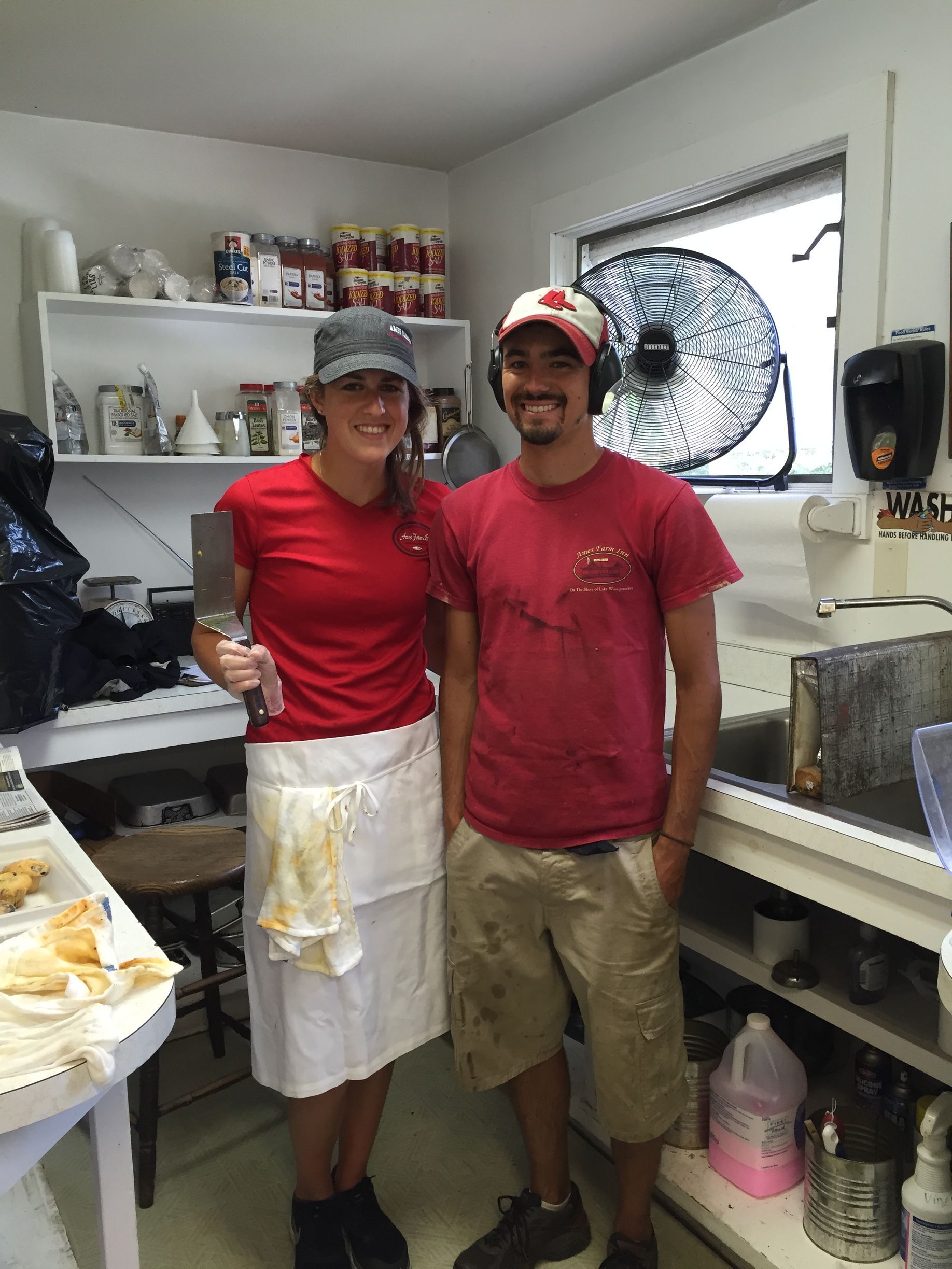 A smiling person in a red shirt and apron holding a cleaver stands beside another person in a red shirt in a kitchen.
