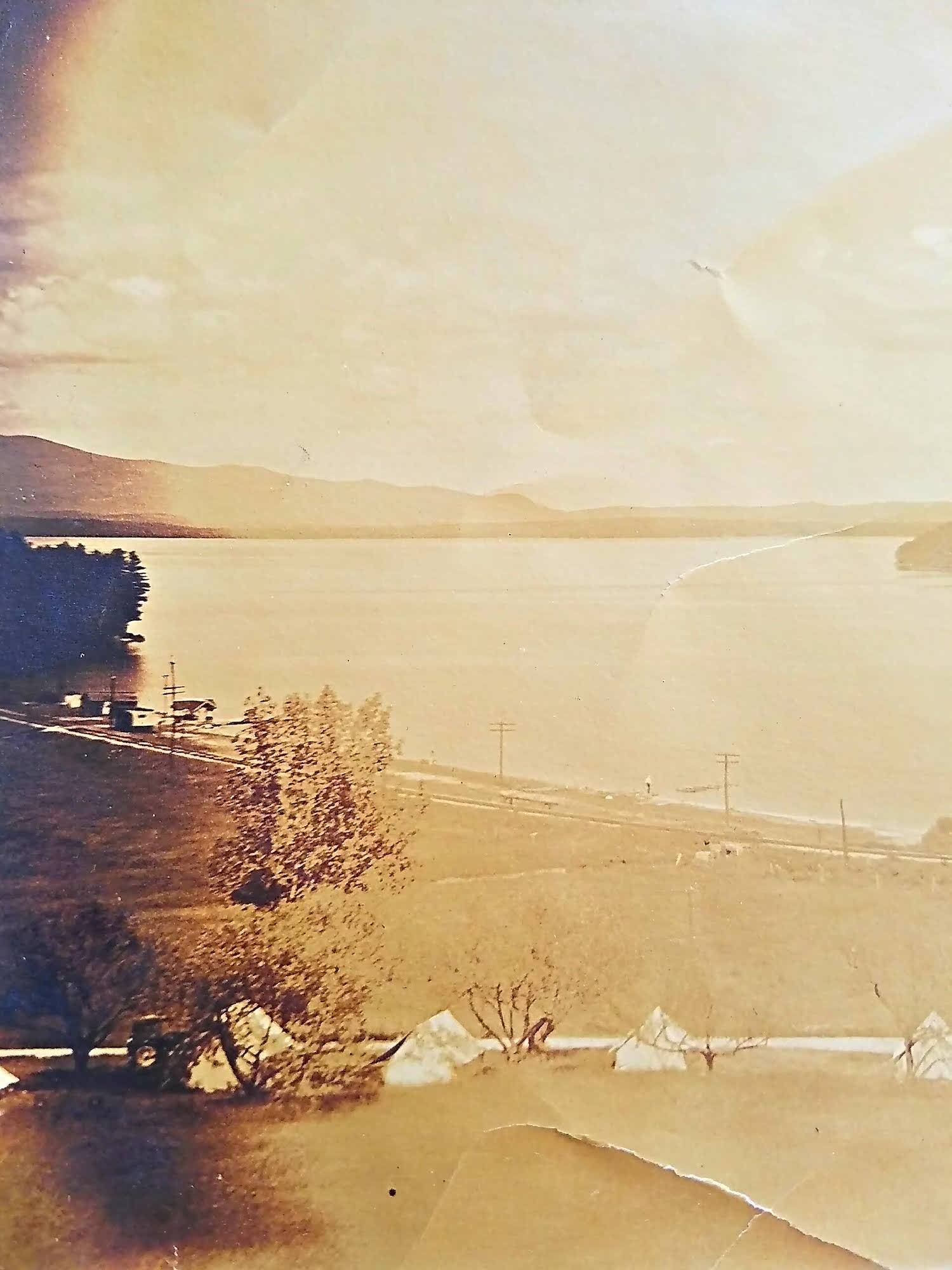 A sepia-toned vintage photograph of tents lined along a lakeshore, with mountains in the distance under a hazy sky.