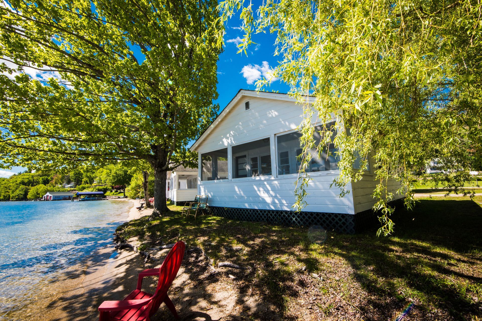 A white cottage with a screened porch sits on a lakeshore next to a large tree with a bright red chair on the sandy beach.