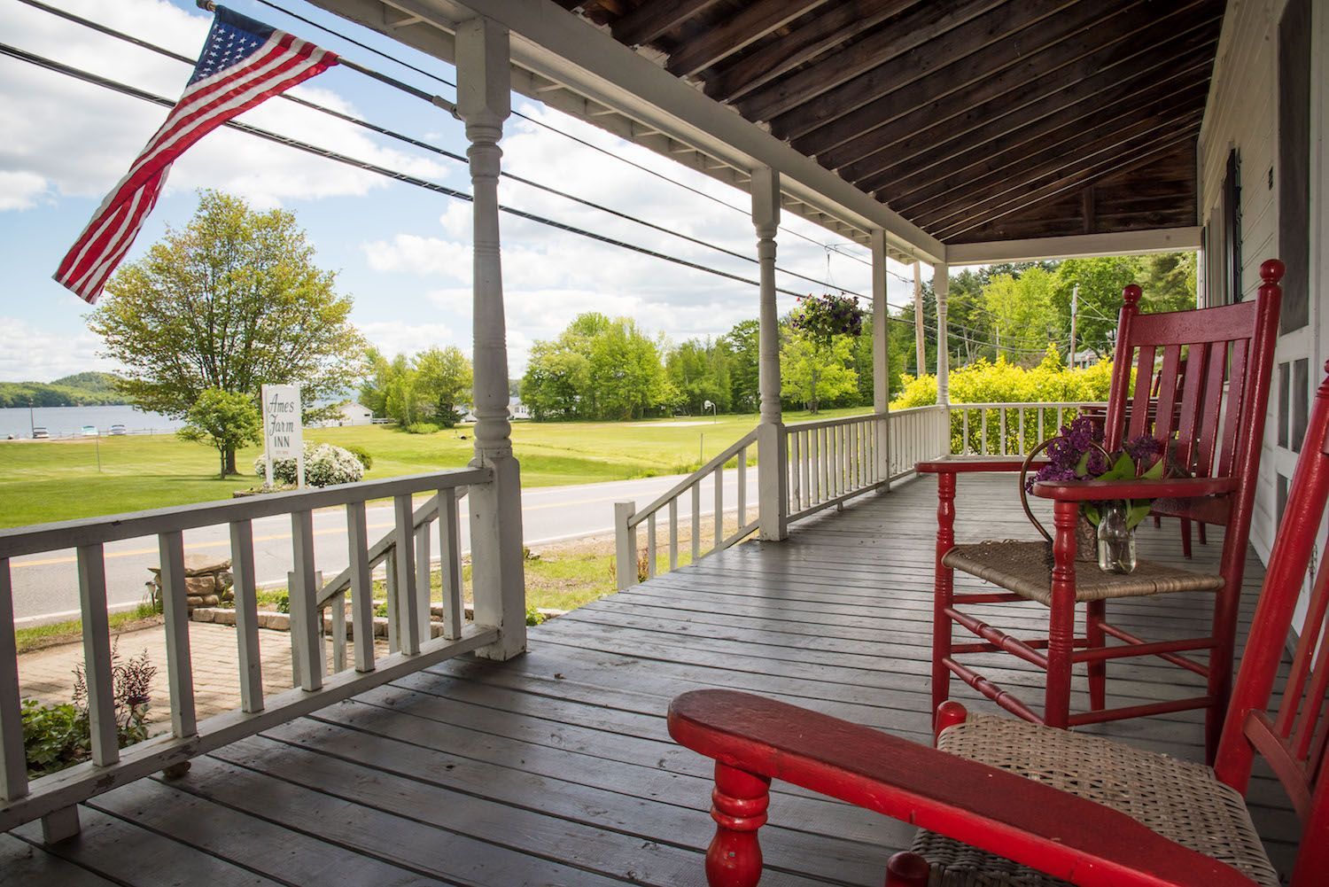 Red rocking chairs on a wood porch overlooking a grassy lawn, a tree-lined lake, and an American flag.