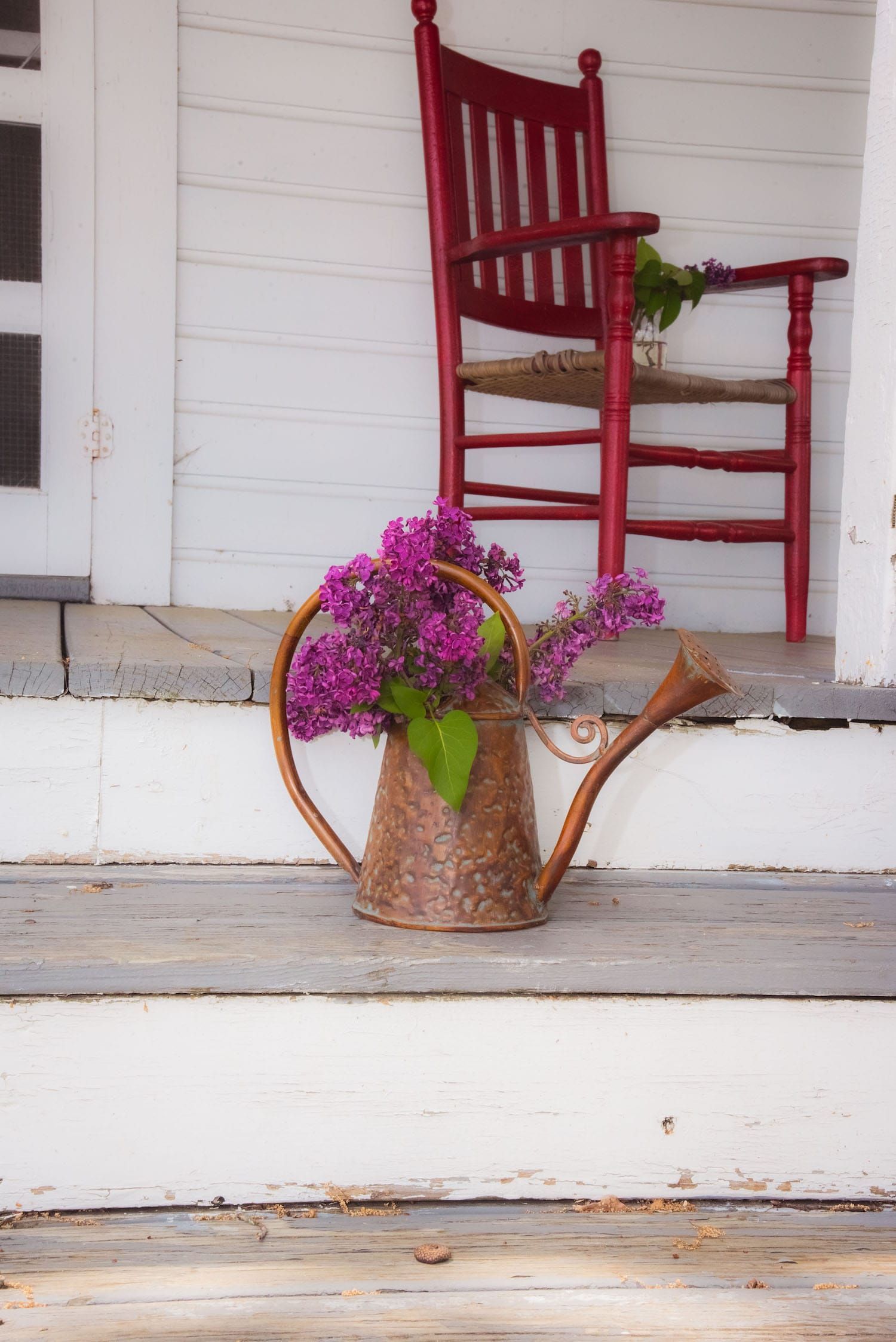 A copper watering can filled with purple lilacs sits on a white porch step in front of a red wooden chair.
