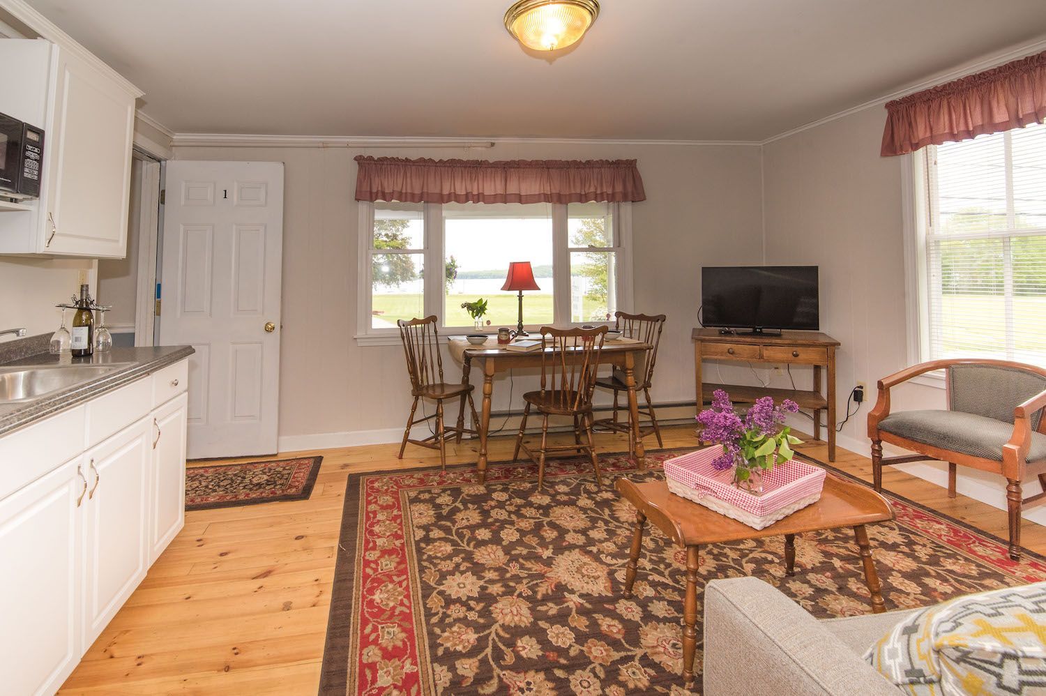 A bright living and dining area with light wood floors, a patterned rug, a kitchen counter, and a window with red trim.