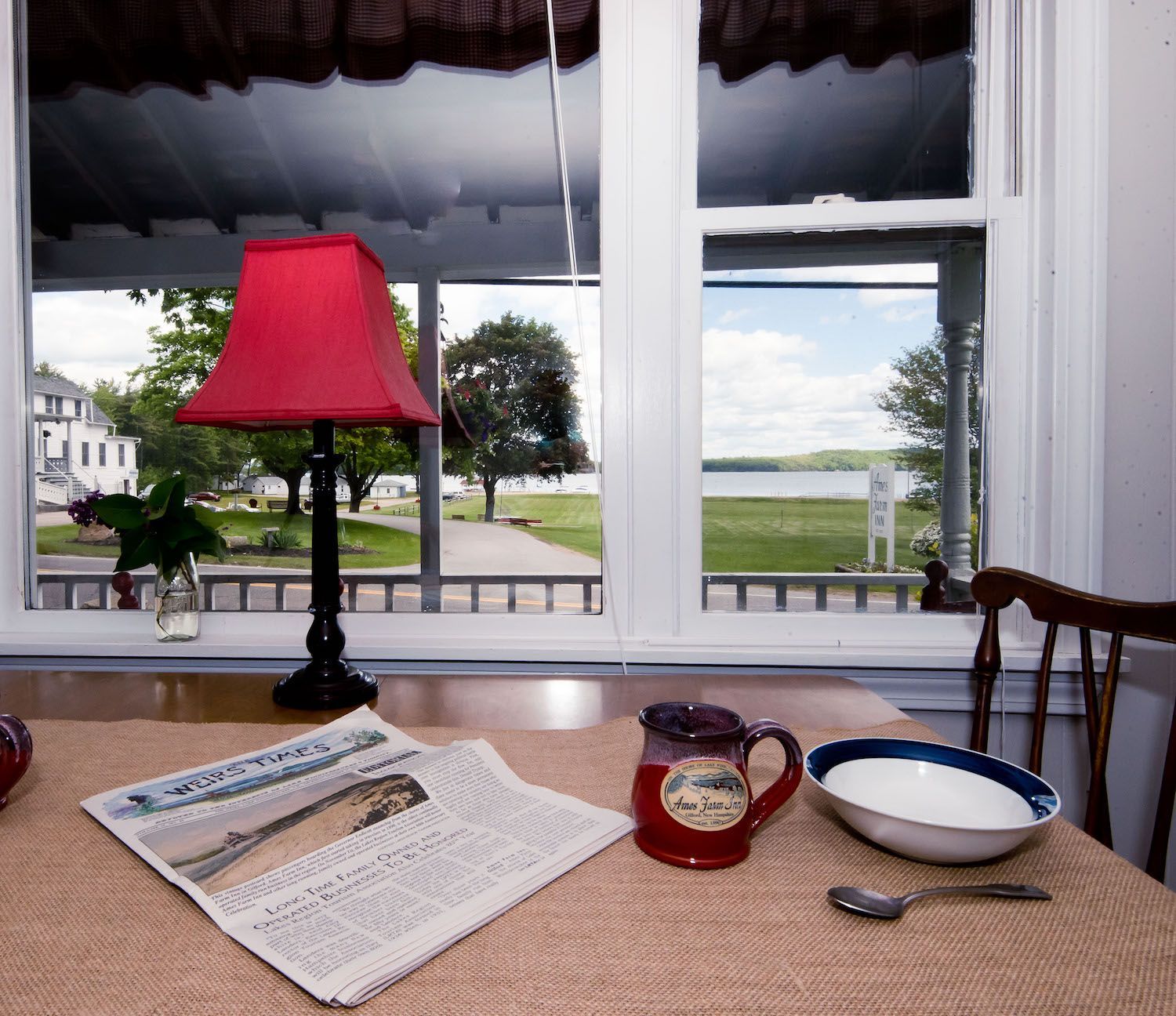 A cozy dining table with a red lamp, a newspaper, a mug, and a bowl, set before a window overlooking a grassy waterfront.