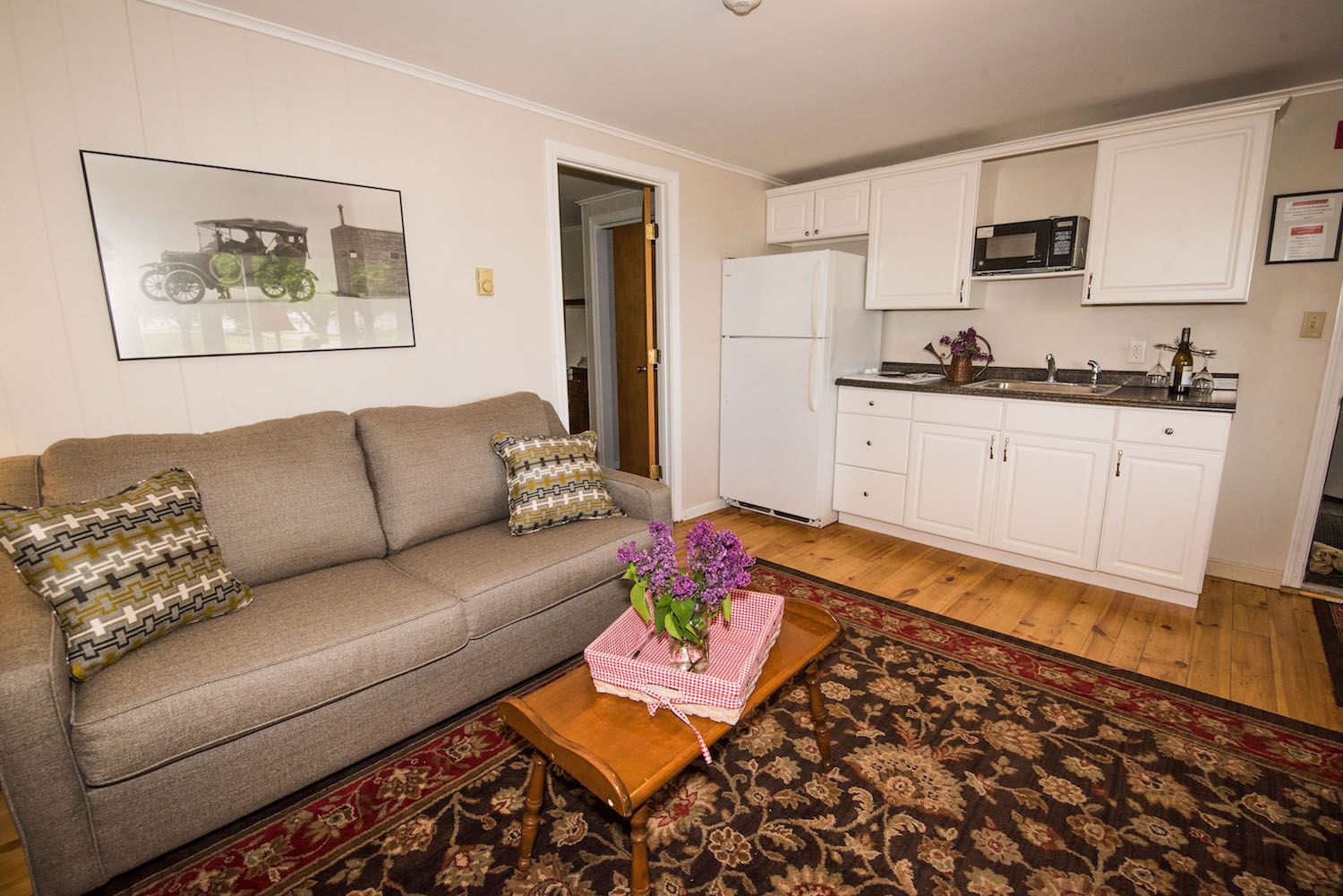 A small living space featuring a gray couch, coffee table with purple flowers, and a kitchenette with white cabinets.