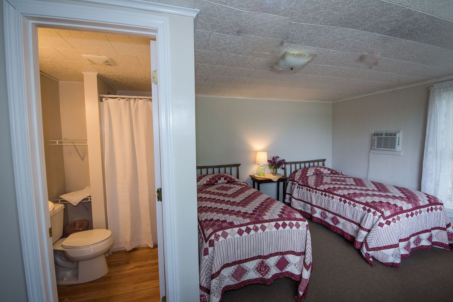 A bedroom with two beds covered in patterned red and white quilts, viewed through an open doorway showing a bathroom.