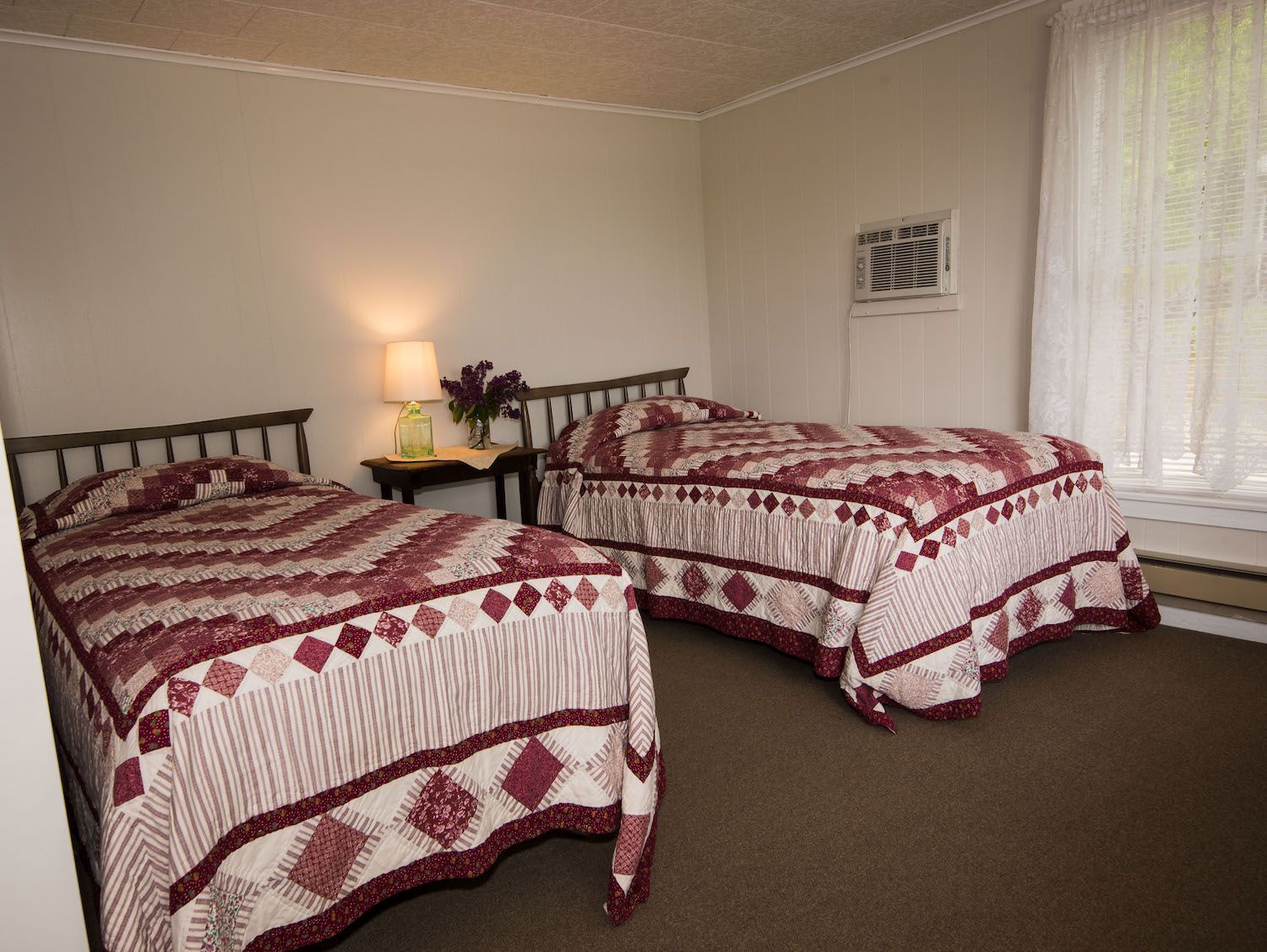 A hotel room with two twin beds featuring maroon and white patchwork quilts, a nightstand with a lamp, and a window.
