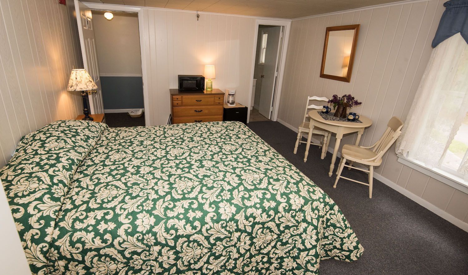 A hotel room features a bed with a green and white patterned quilt, a small dining table with two chairs, and a dresser.