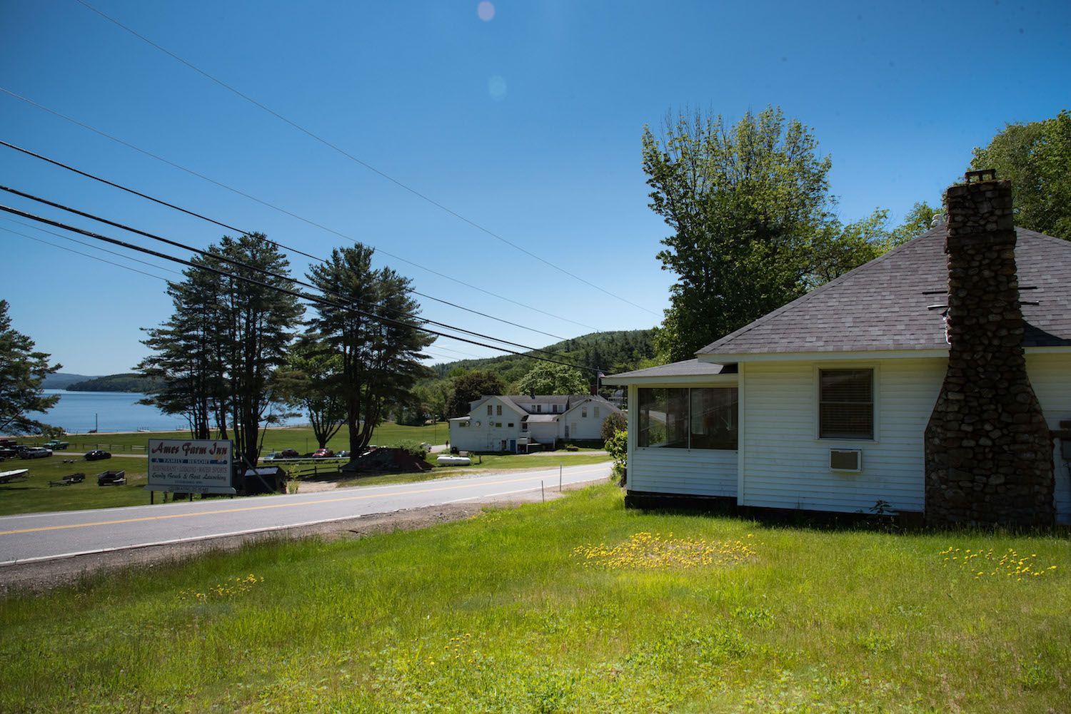A white cottage with a stone chimney sits beside a road near a lake on a sunny day with trees and blue sky.