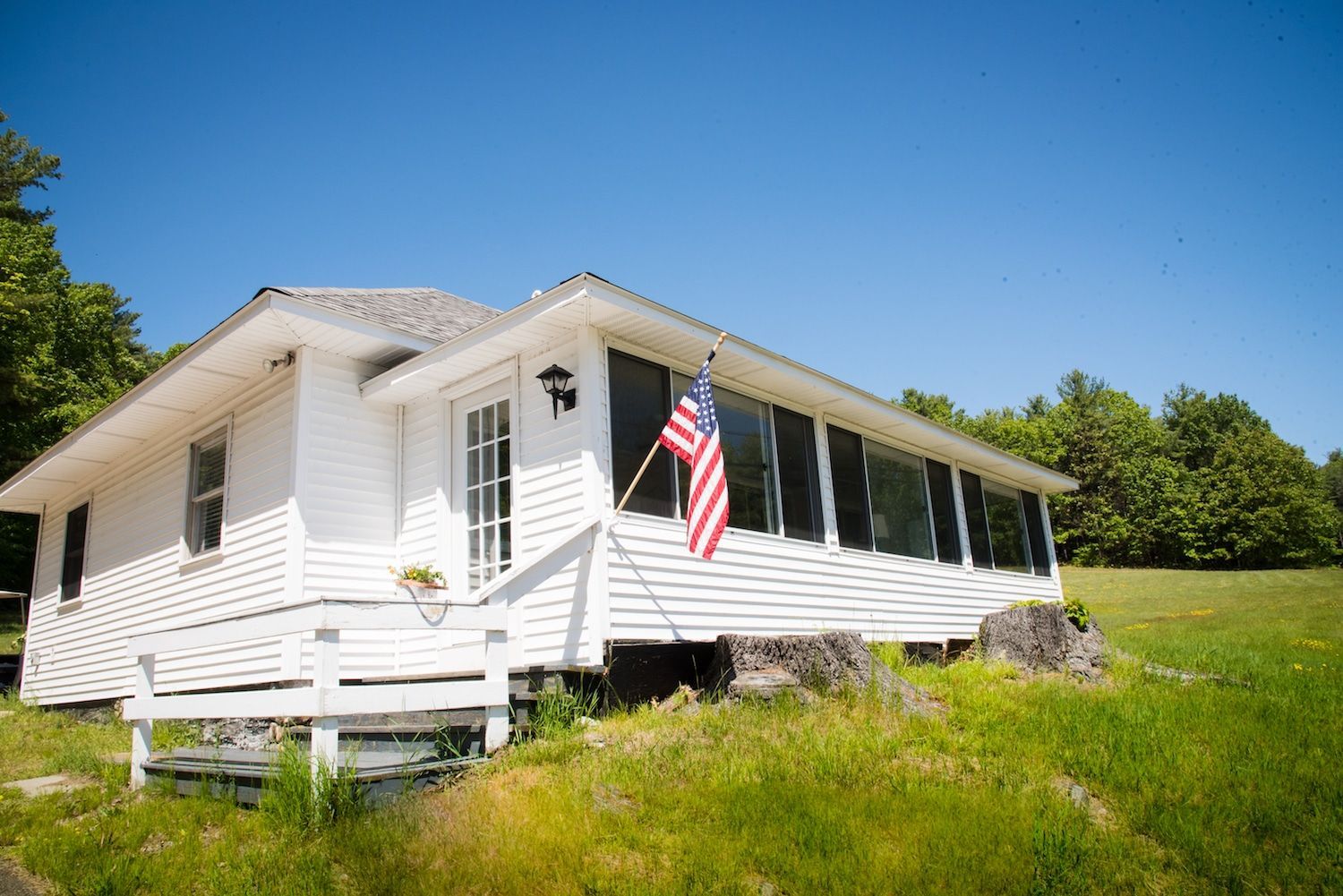 A white cottage sits on a grassy hill under a bright blue sky, with an American flag displayed near the front porch.