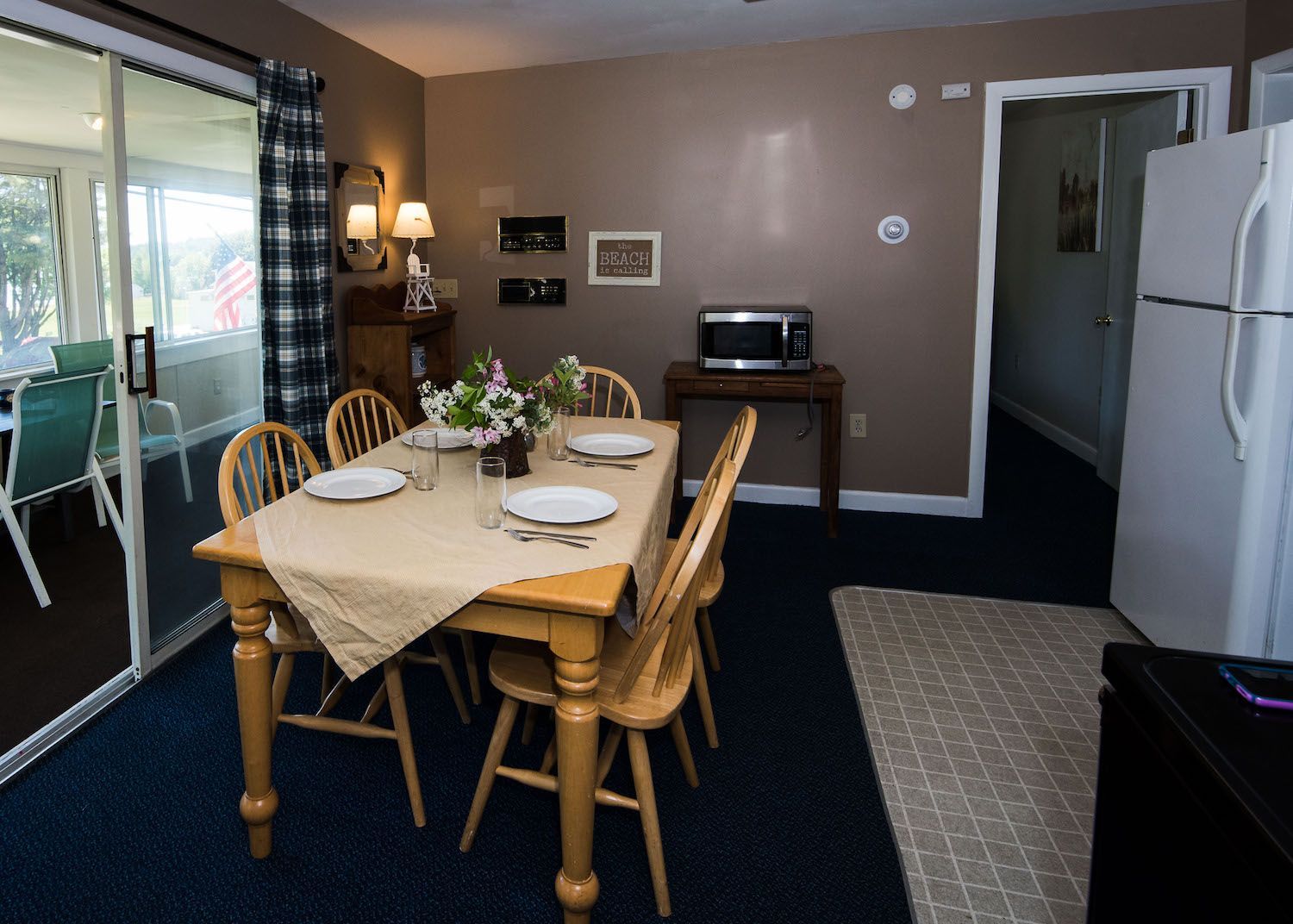 A dining set in a room with brown walls and blue carpet, including a table, four chairs, a microwave, and a refrigerator.