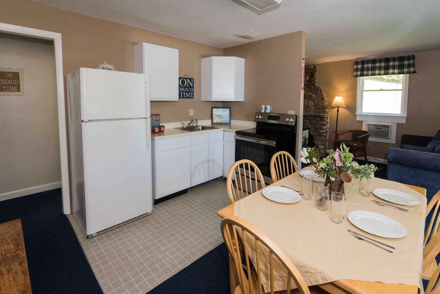 A kitchen area with a white refrigerator, cabinets, stove, and a dining table with chairs in a carpeted room.
