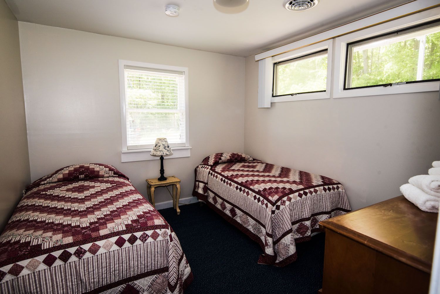 A bedroom featuring two twin beds with matching burgundy patterned quilts, a small nightstand with a lamp, and white walls.