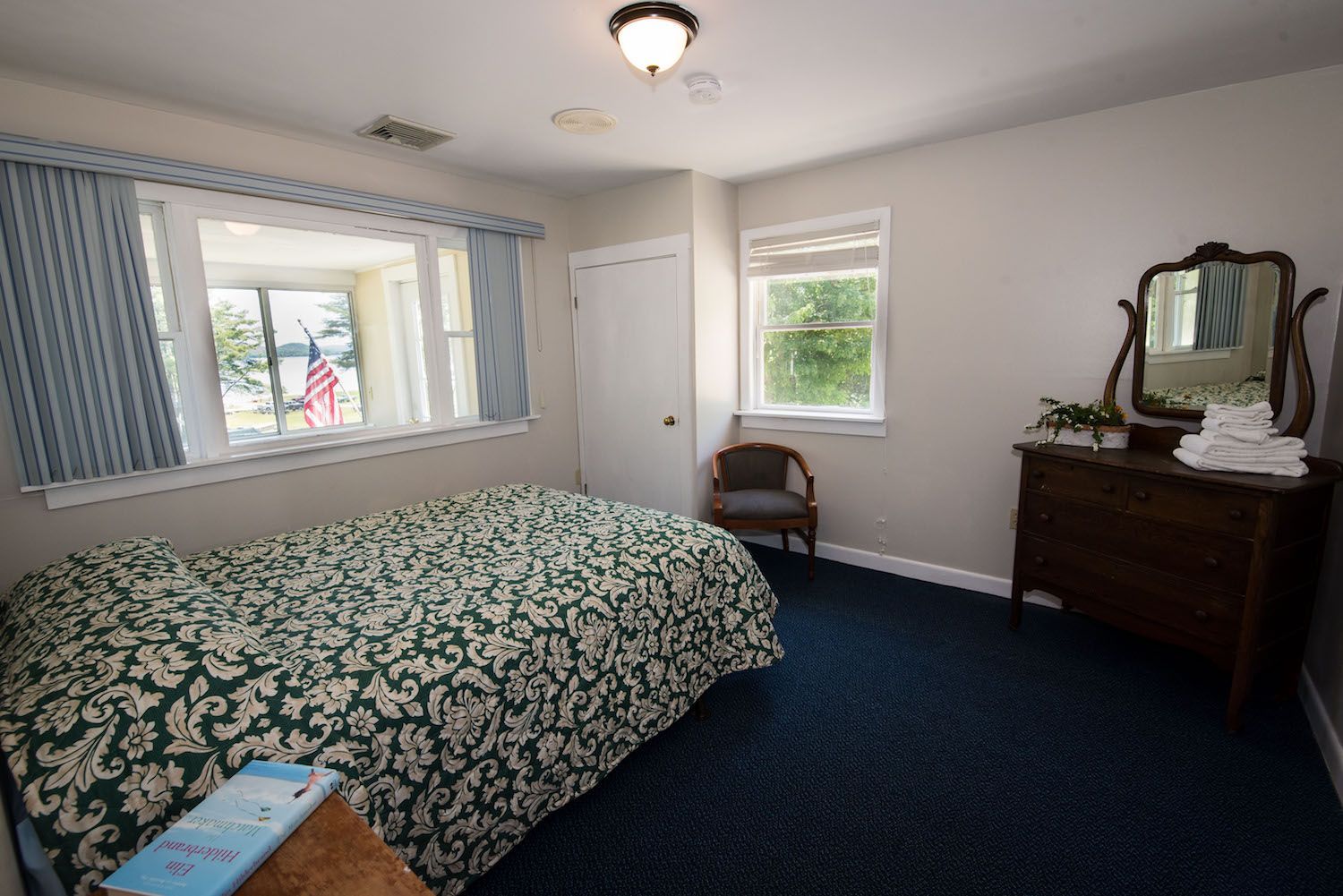 A hotel room with a patterned bedspread, dark blue carpet, a wooden dresser with a mirror, and a window with curtains.