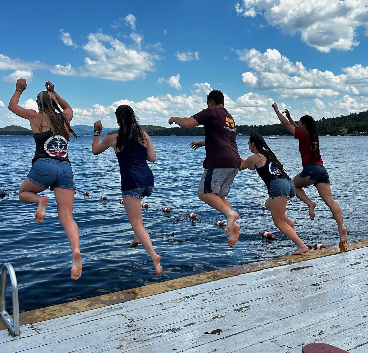 Five people leap from a white wooden dock into a blue lake under a bright, sunny sky with scattered clouds.