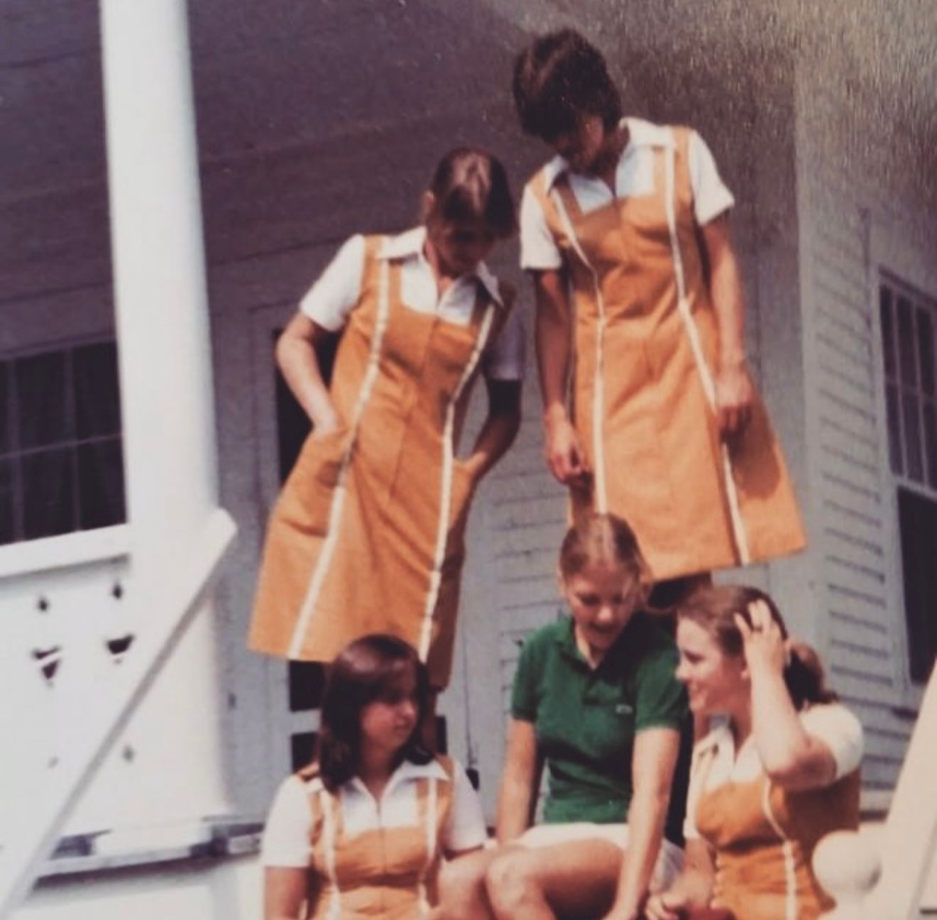 A group of five people on a porch, four wearing matching orange jumper dresses with white collared shirts, one in green.
