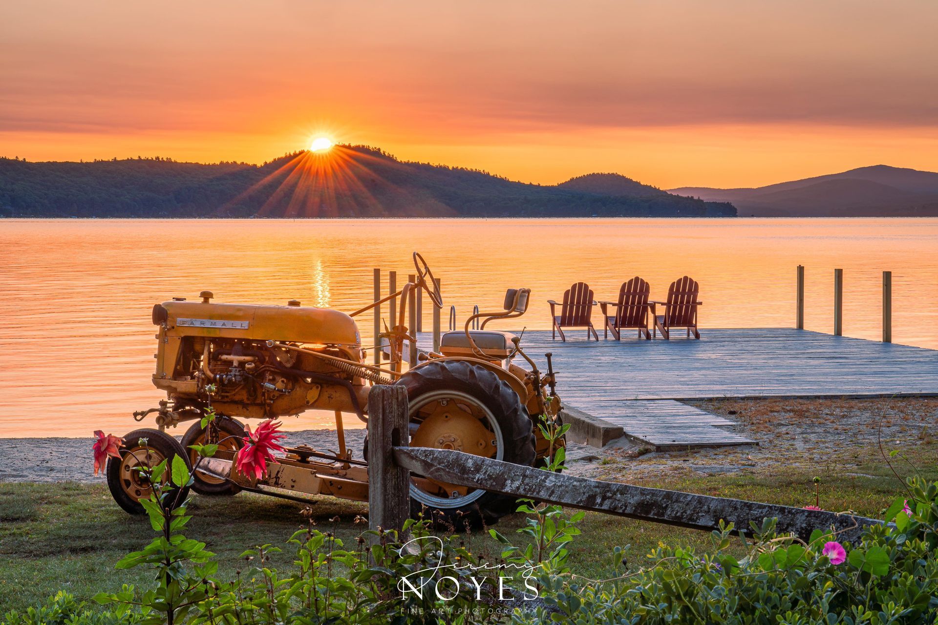 A golden vintage tractor sits on a grassy shore beside a wooden dock with three chairs at sunset over a calm lake.