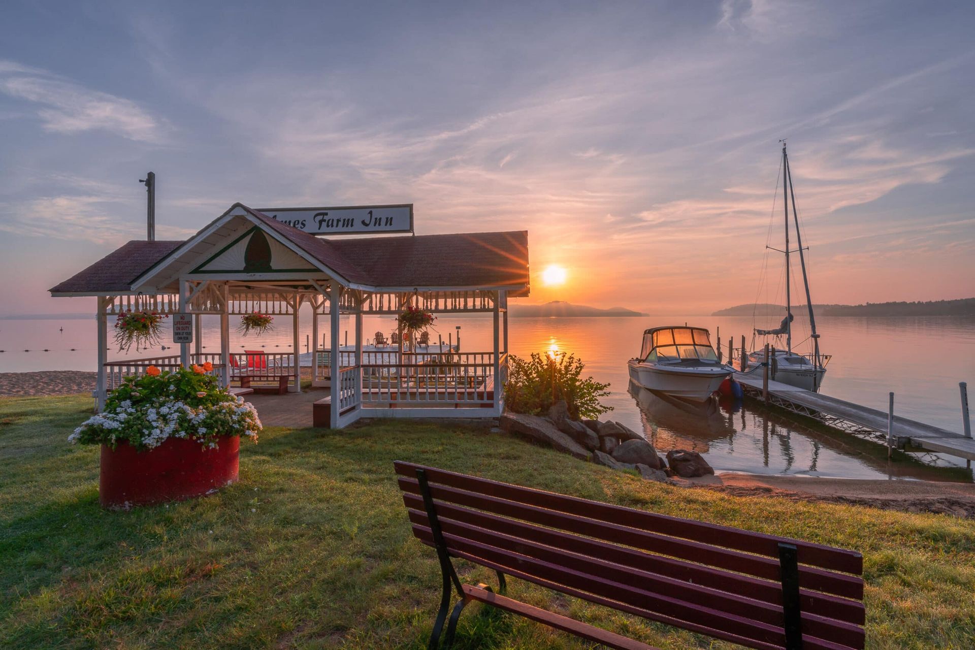 A wooden bench faces a gazebo, boats at a dock, and a calm lake under a colorful sunset sky.