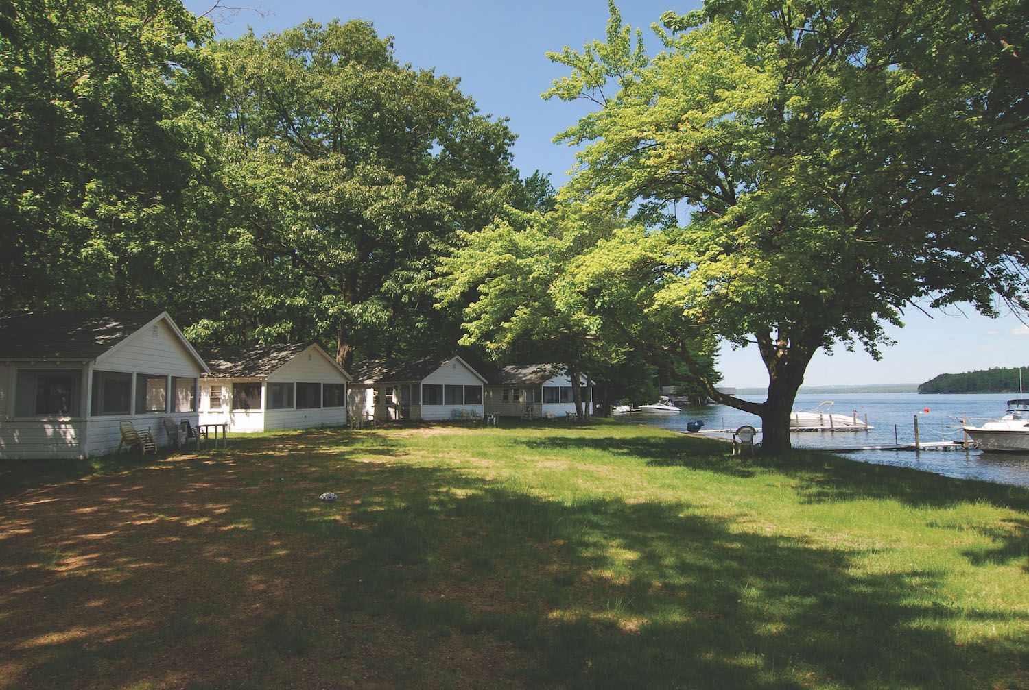 A row of white lakeside cottages nestled among lush trees next to a green grassy lawn and a blue lake.