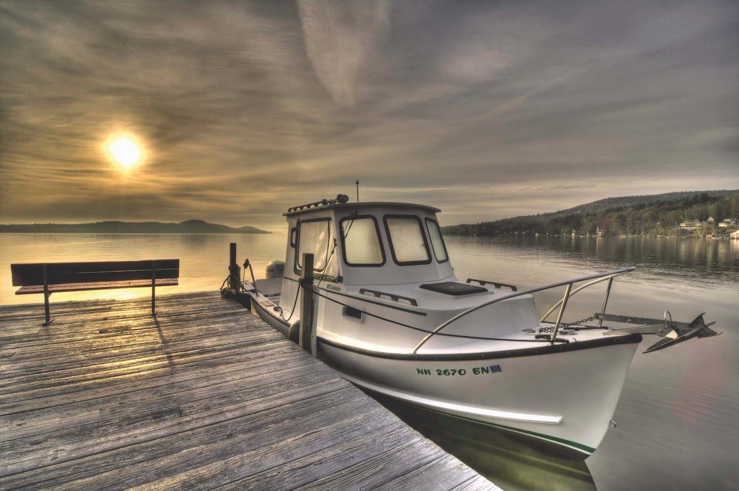A white motorboat tied to a wooden dock at sunset on a calm lake, with distant hills under a moody, cloudy sky.