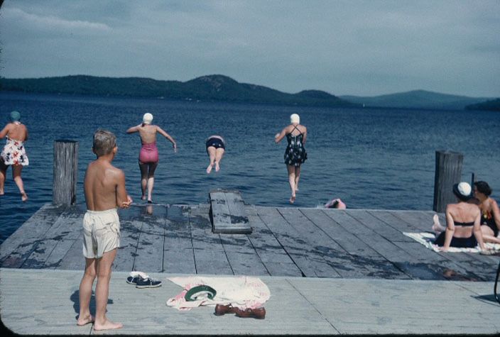 People in swimsuits jump off a wooden dock into a large lake with mountains in the background.