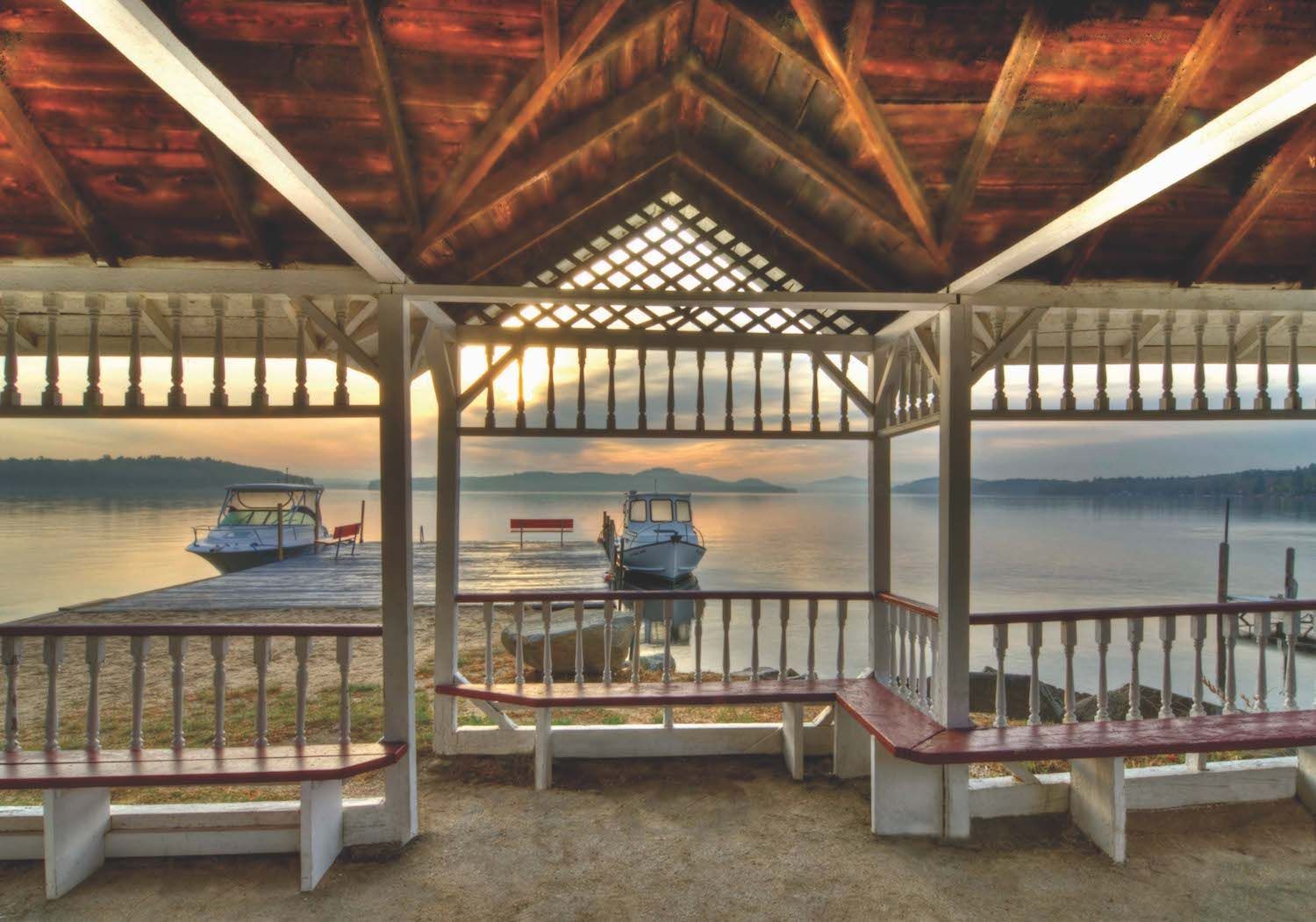 A gazebo overlooks a calm lake at sunset, with two small motorboats docked nearby under a warm, glowing sky.