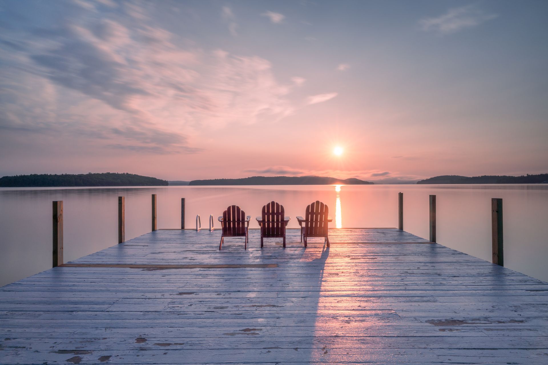 Three red Adirondack chairs sit on a weathered wooden dock facing a calm lake under a soft, golden sunrise.