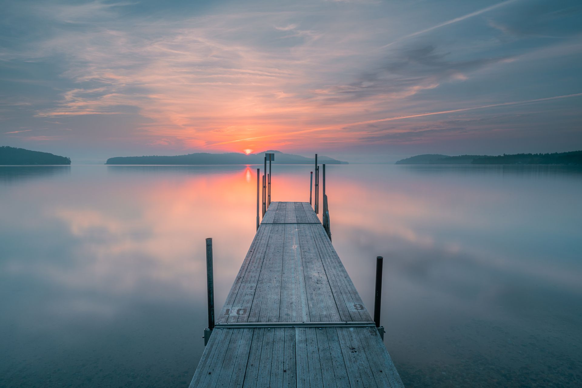 A wooden dock extends into a calm lake at sunrise, with soft pink and orange light reflecting on the still water.