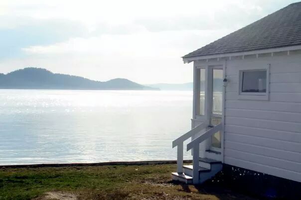 A white cabin with steps overlooks a calm, reflective body of water with distant hills under a bright sky.