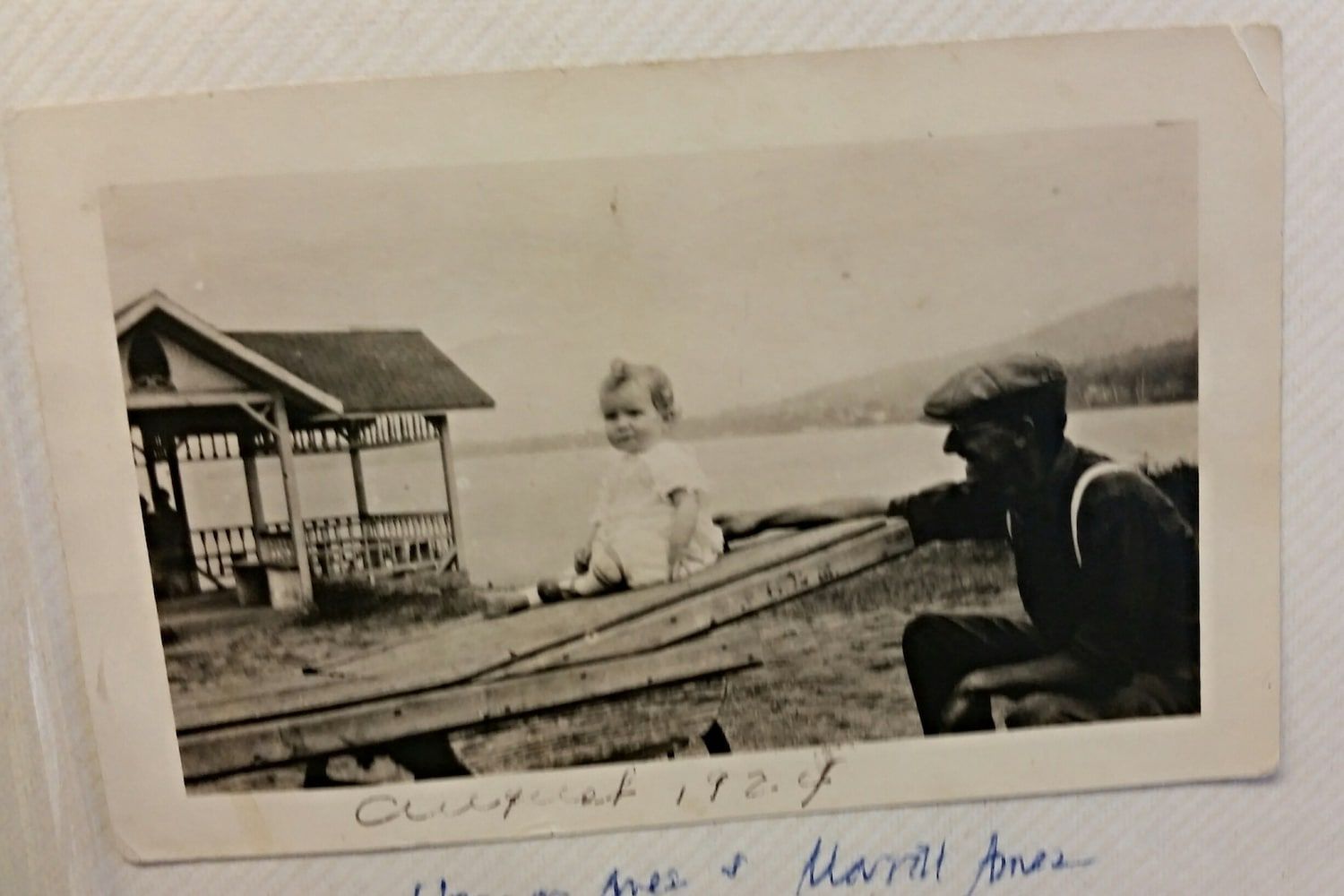 A vintage photo shows a small child sitting on a wooden board while an adult kneels nearby by a lake with a gazebo.