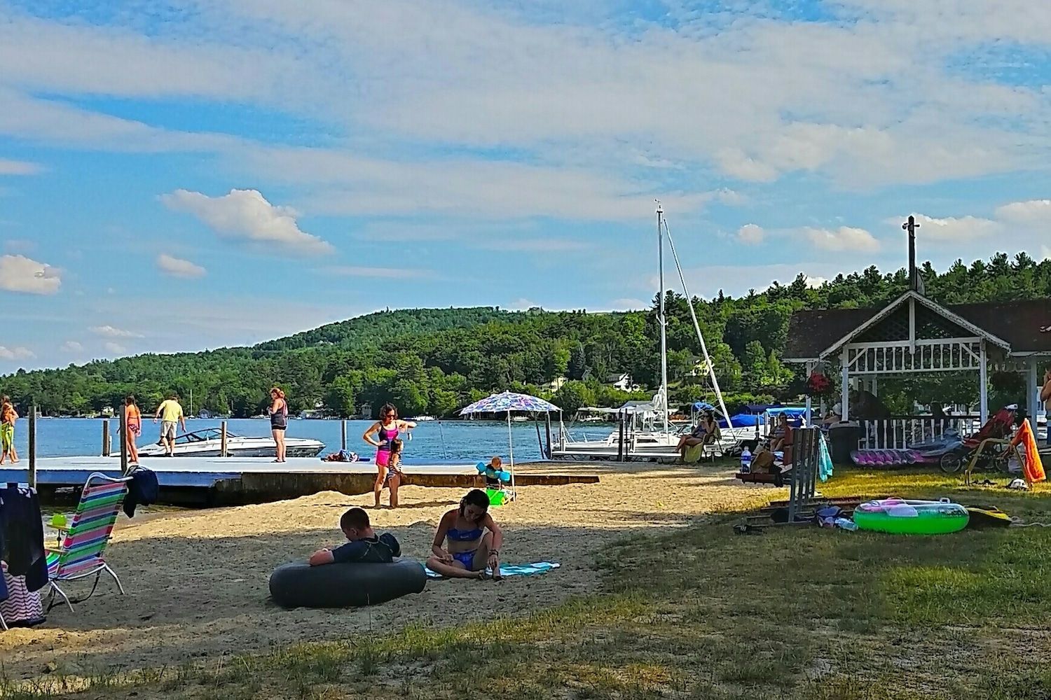 People relaxing on a sandy beach by a lake with a wooden dock, boats, and a gazebo on a sunny day.