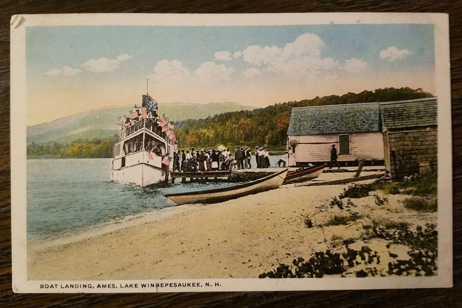 A vintage colorized postcard of a steamship docked at a shore in Lake George, N.Y., with a small boat and wooden building.
