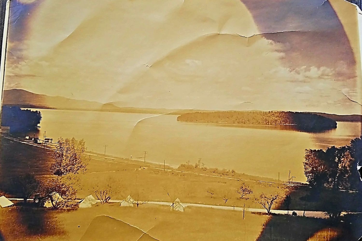 A sepia-toned vintage photograph of a large lake with a grassy shoreline, distant hills, and small white tents in the grass.