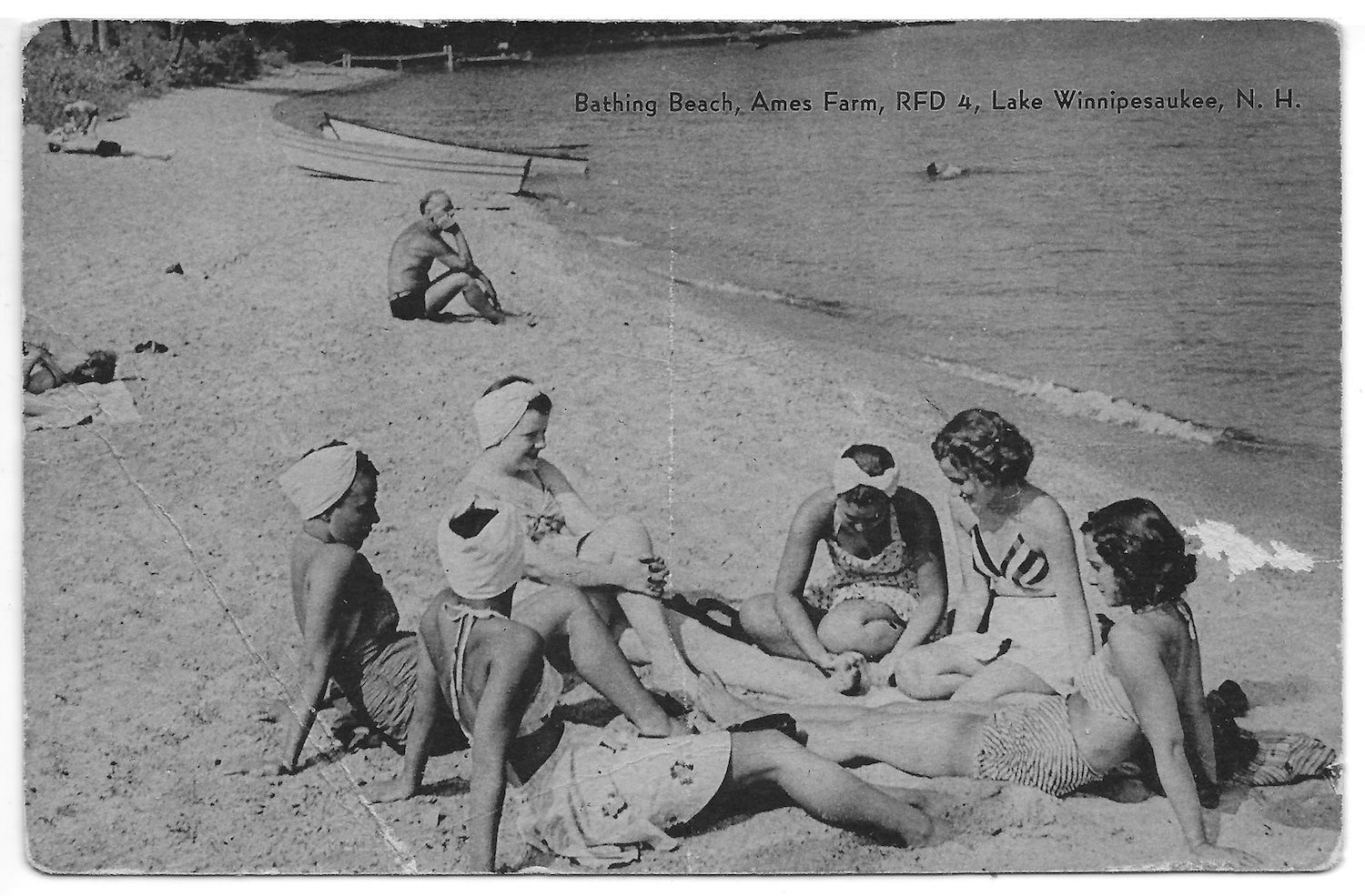 A group of people in vintage swimwear sitting on a sandy beach by a lake, with one person sitting alone in the distance.