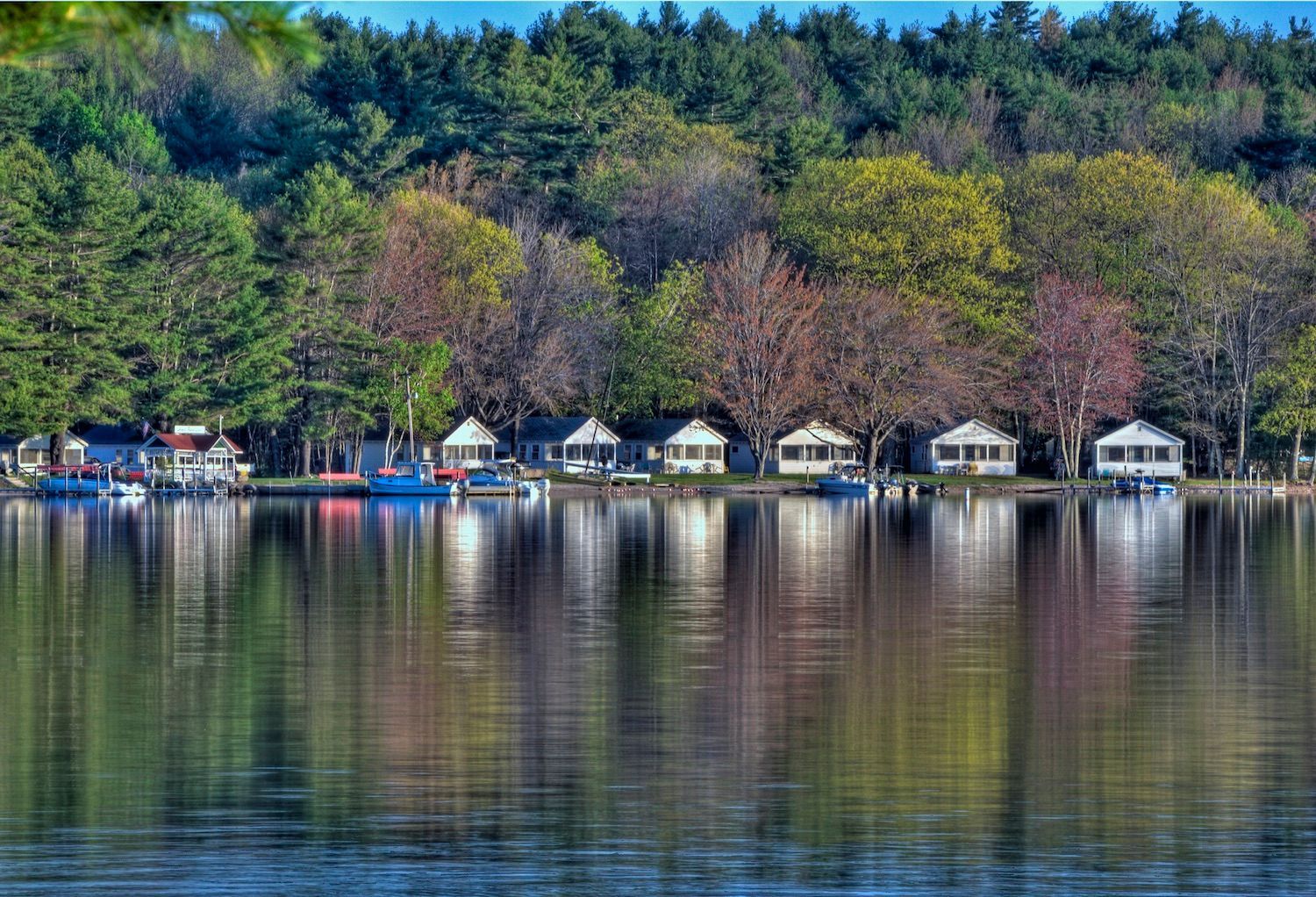 A row of small white cabins lines the shore of a calm lake, reflected in the water with a backdrop of green and fall trees.