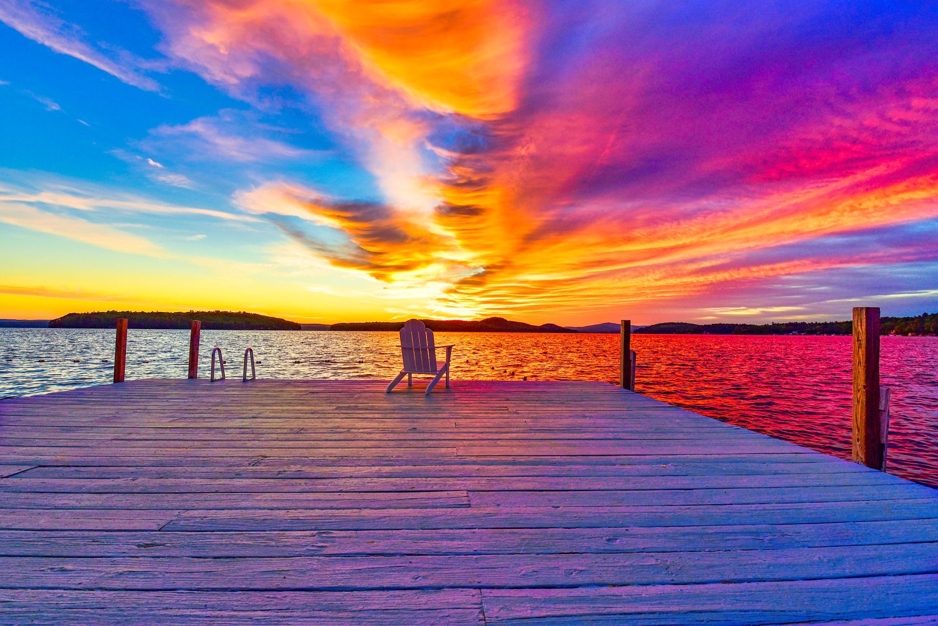 A lone chair sits on a wooden dock overlooking a vibrant, colorful sunset over the water.