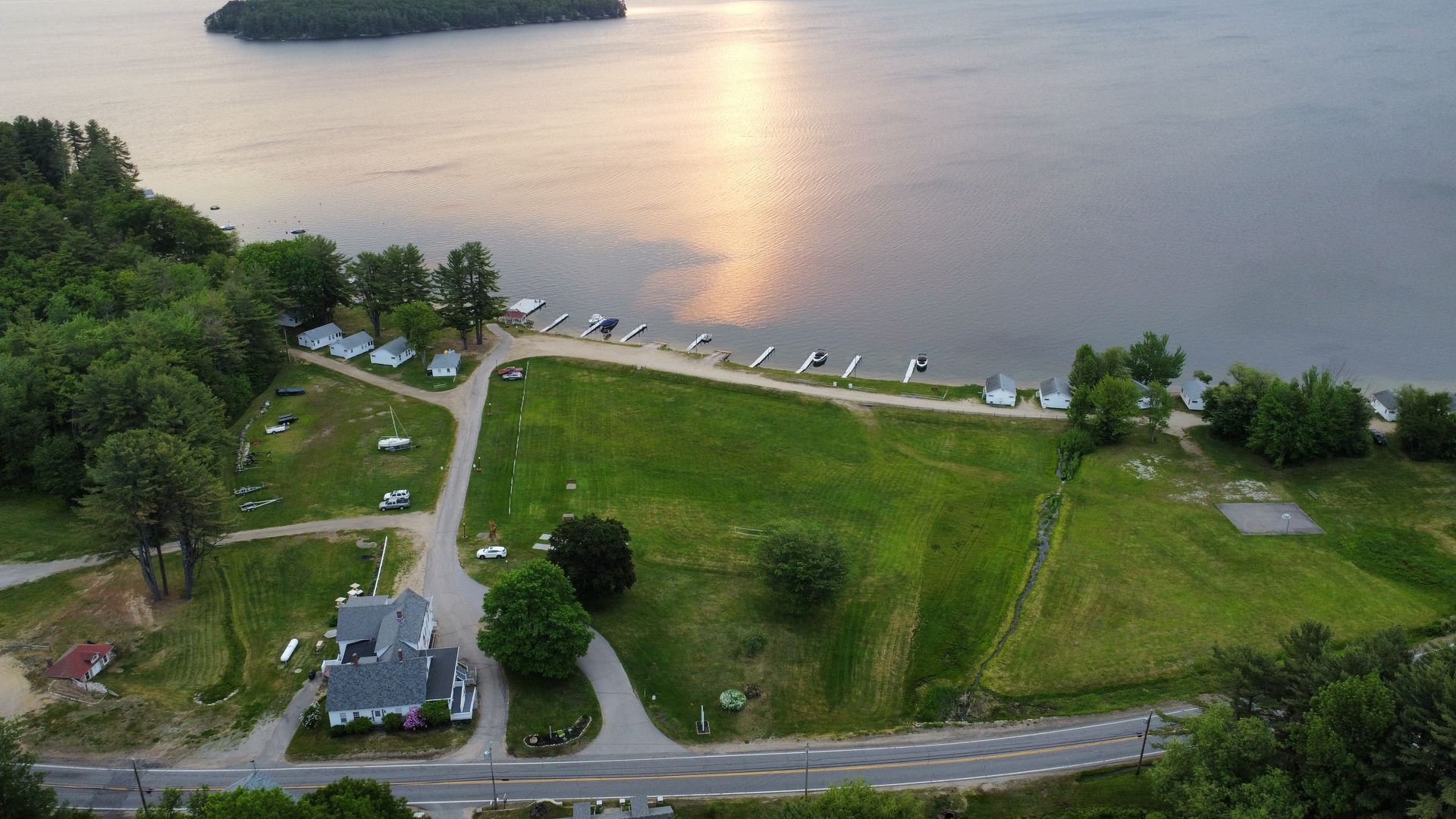 Aerial view of a grassy campground with a house, gravel paths, and boat docks along a lake shore at sunset.