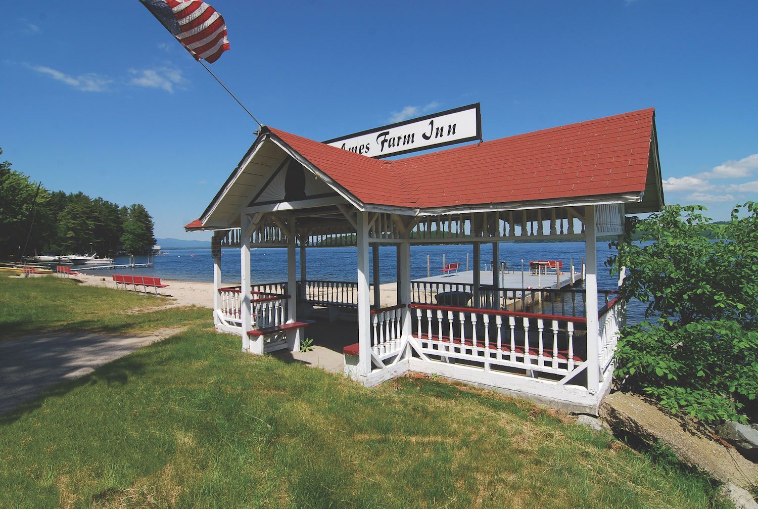 A white and red gazebo with a red roof sits on a grassy area overlooking a lake.