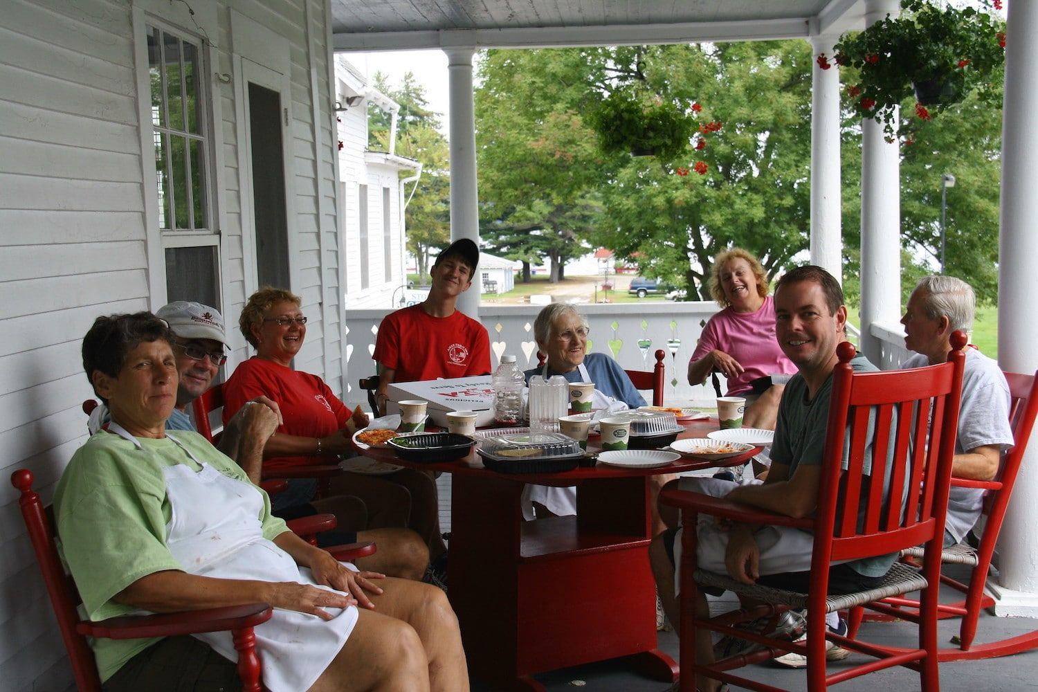 A group of people sit around a table on a white porch, enjoying a meal together in red rocking chairs.