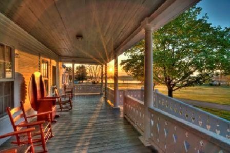 A sunlit wooden porch with several red rocking chairs overlooks a grassy lawn and trees at sunset.
