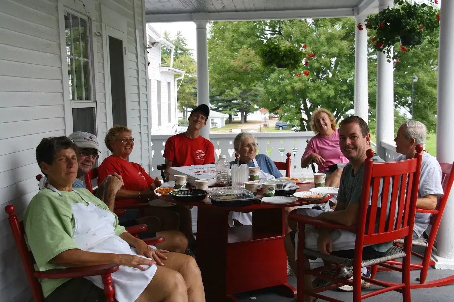 A group of people sit around a table on a white porch, enjoying a meal together in red rocking chairs.