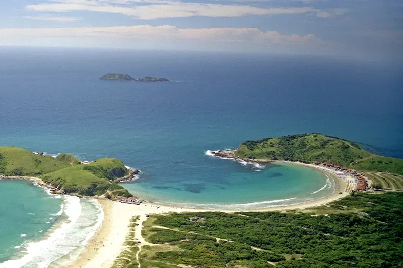 Vista aérea de uma baía com água turquesa, praias de areia e colinas verdejantes sob um céu claro.