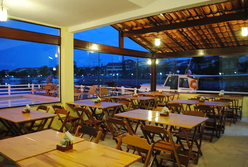 Interior de restaurante com mesas e cadeiras de madeira, grandes janelas com vista para a água, à noite.