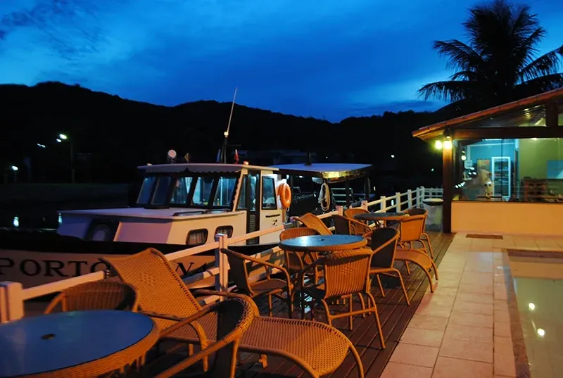 Barco atracado em um restaurante à beira-mar com mesas e cadeiras ao entardecer. Montanhas e céu azul escuro ao fundo.