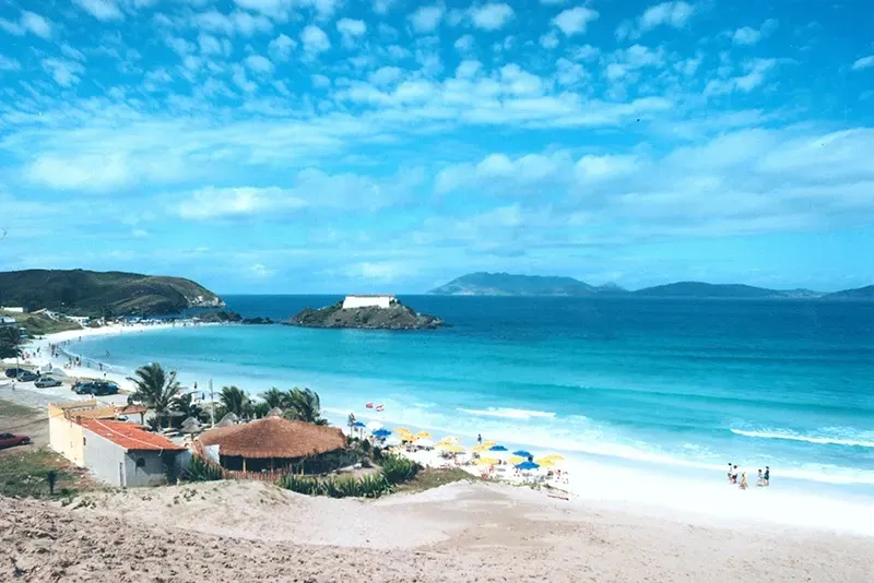 Cena de praia com areia branca, água turquesa, céu azul e um pequeno forte ao longe.