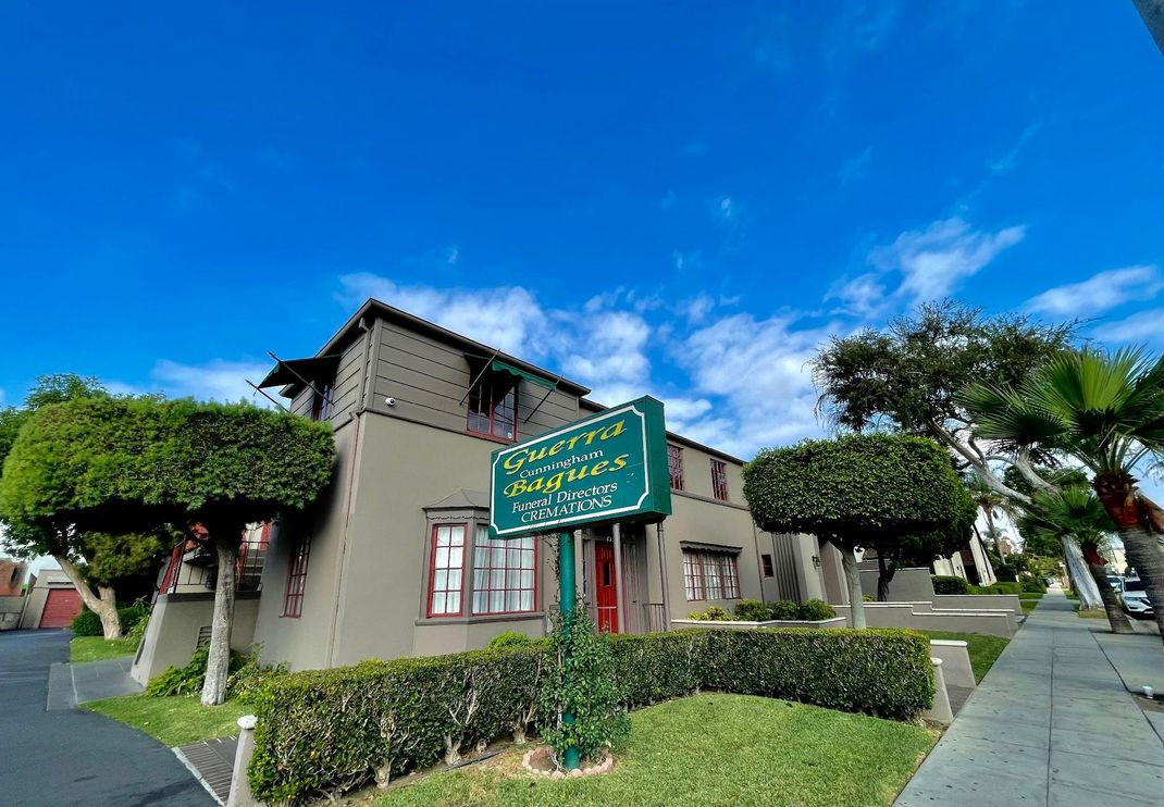 A two-story tan building with a sign that reads "Gardenia Apartments" under a bright blue sky with scattered clouds.